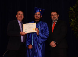 Graduate in blue gown holding diploma with officials