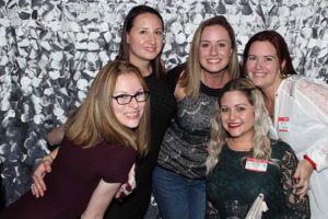 Five women smiling together against silver backdrop