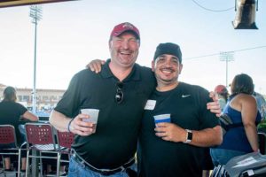Two men smiling, holding drinks at baseball stadium