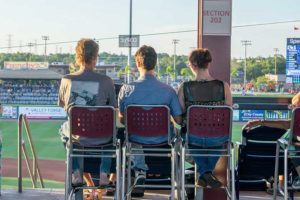 Three people seated in stadium watching baseball